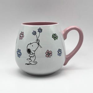 May include: White ceramic mug featuring Snoopy holding flowers, with a pink handle and interior. The mug is decorated with colorful floral accents. A charming and whimsical design for coffee or tea.