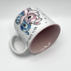 May include: White ceramic mug featuring a blue and pink cartoon character design. The mug has a pink interior and a white handle. The words "We're better together" are printed on the side of the mug.