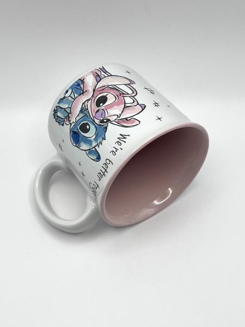 May include: White ceramic mug featuring a blue and pink cartoon character design. The mug has a pink interior and a white handle. The words "We're better together" are printed on the side of the mug.
