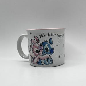 May include: White ceramic mug featuring a blue and pink cartoon character hugging. The mug has a pink rim and the text "We're better together" is printed on the side. The mug has a white handle.