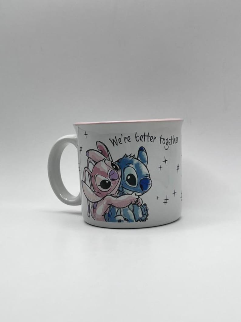 May include: White ceramic mug featuring a blue and pink cartoon character hugging. The mug has a pink rim and the text "We're better together" is printed on the side. The mug has a white handle.
