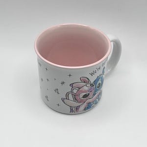May include: White ceramic mug with a pink interior and handle. The mug features a colorful illustration of two cartoon characters embracing, with the words "We're better" visible. Small crosses and hash marks are also printed on the mug.