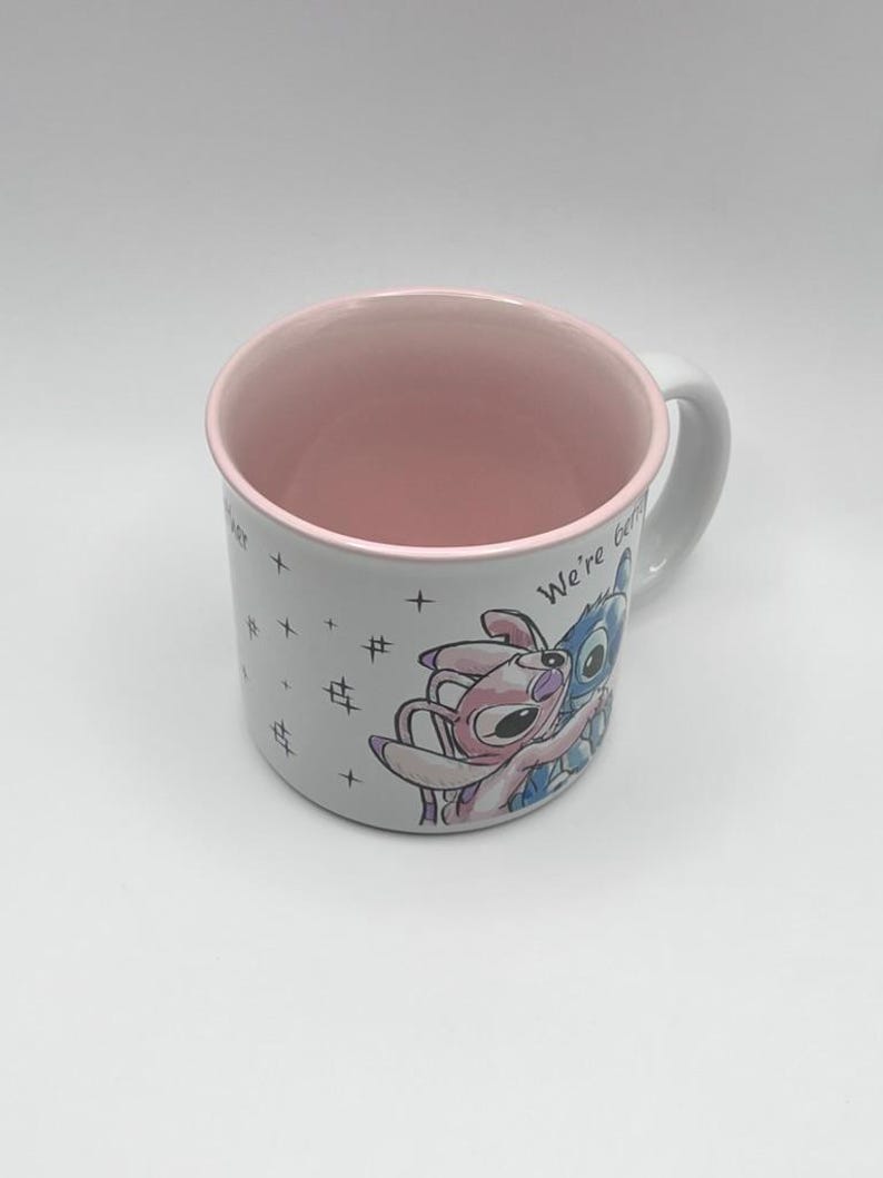 May include: White ceramic mug with a pink interior and handle. The mug features a colorful illustration of two cartoon characters embracing, with the words "We're better" visible. Small crosses and hash marks are also printed on the mug.
