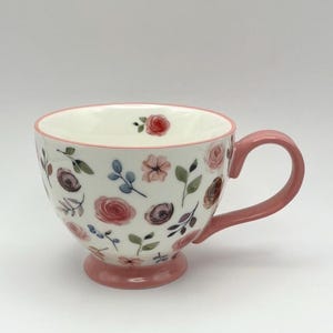 Può includere: Una tazza in ceramica bianca con un delicato motivo floreale ad acquerello con fiori e foglie rosa, viola e verdi. La tazza presenta un bordo rosa, un manico rosa e una base a piedistallo rosa, con una piccola rosa dipinta sul bordo interno.