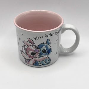 May include: White ceramic mug with a pink interior and a gray handle. The mug features a cartoon illustration of two characters embracing, with the text "We're better together". The mug is decorated with small hash marks.