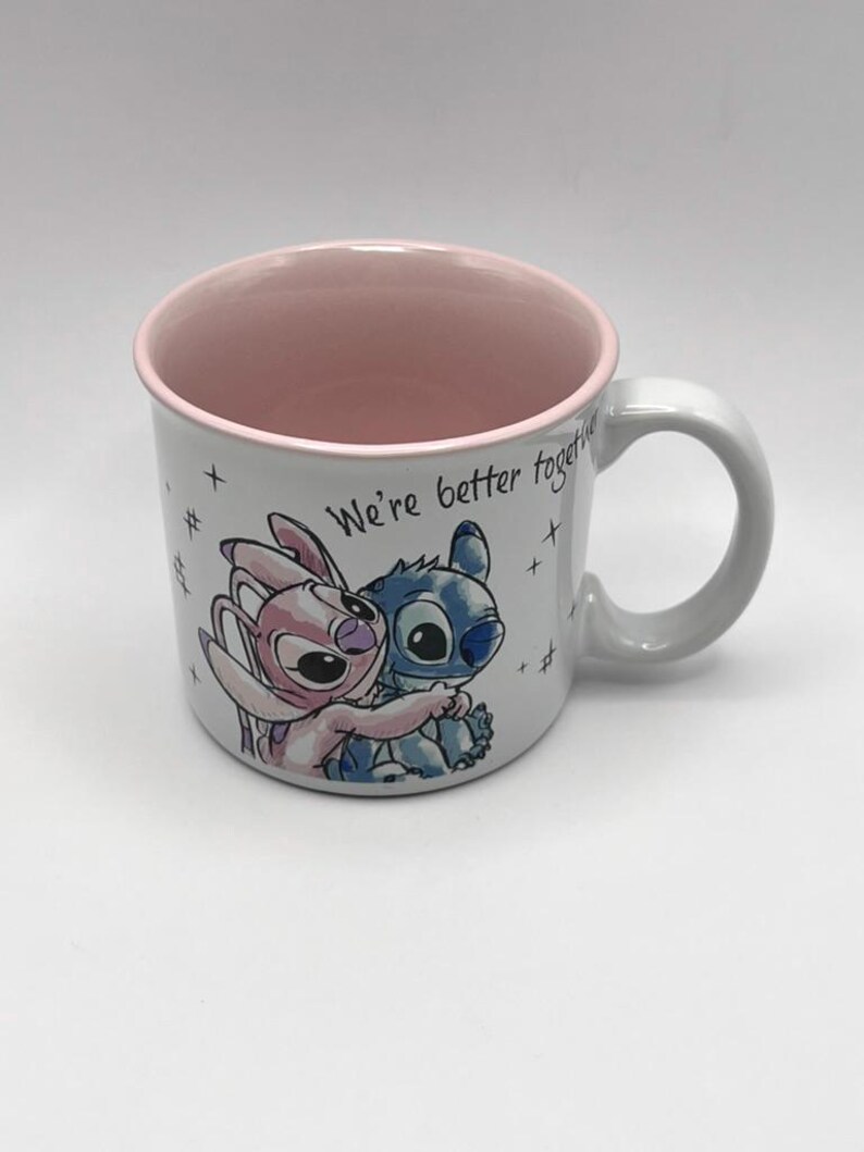 May include: White ceramic mug with a pink interior and a gray handle. The mug features a cartoon illustration of two characters embracing, with the text "We're better together". The mug is decorated with small hash marks.