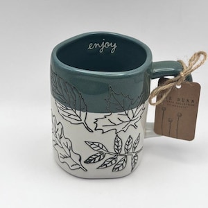 May include: A ceramic mug with a teal top and white bottom, featuring black leaf illustrations. The word "enjoy" is written inside the mug. A tag is attached to the handle with twine.