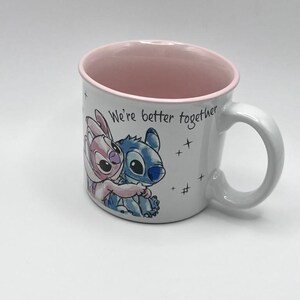 May include: White ceramic mug featuring a cartoon image of Stitch and Angel hugging, with the text "We're better together." The interior and rim of the mug are pink, and the handle is white. Small star graphics are scattered around the text.