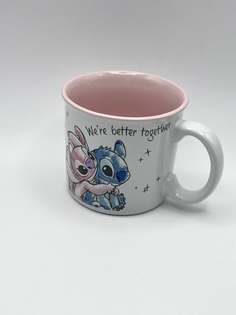 May include: White ceramic mug featuring a cartoon image of Stitch and Angel hugging, with the text "We're better together." The interior and rim of the mug are pink, and the handle is white. Small star graphics are scattered around the text.