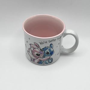 May include: White ceramic mug with a pink interior and handle. The mug features a colorful illustration of two cartoon characters embracing, with the text "We're better together" written on the side. The mug is a standard size.