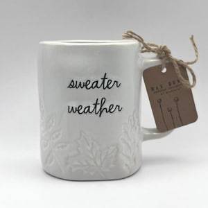 Peut inclure: Tasse en céramique blanche avec l'inscription "sweater weather" en écriture noire. La tasse présente des motifs de feuilles en relief et une étiquette marron attachée avec de la ficelle. L'étiquette indique "RAE DUNN ARTISAN COLLECTION by Magenta."