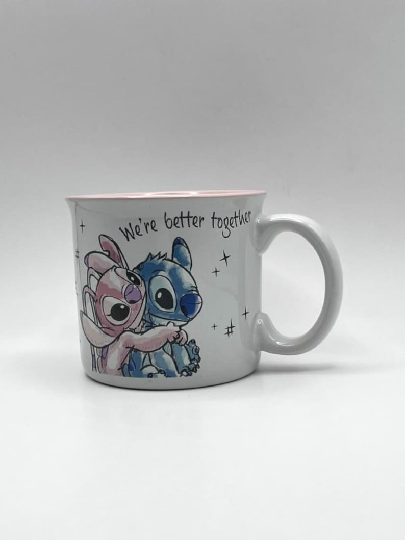May include: A white ceramic mug with a pink interior and a curved handle. The mug features a colorful illustration of two cartoon characters embracing, with the text "We're better together".