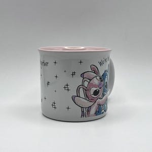 May include: A white ceramic mug with a pink interior rim. The mug features a cartoon illustration of two characters embracing, with the words "We're better together" and star graphics. The mug has a handle on the right side.