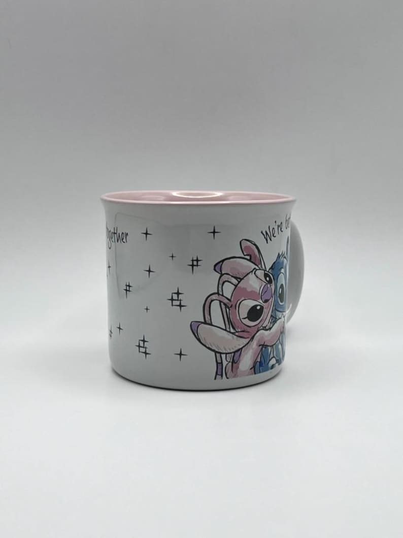 May include: A white ceramic mug with a pink interior rim. The mug features a cartoon illustration of two characters embracing, with the words "We're better together" and star graphics. The mug has a handle on the right side.