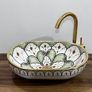 May include: Oval ceramic sink with a floral design in white, green, and red. The sink has a gold-coloured rim and a matching tap. The sink is placed on a wooden surface.