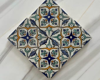 May include: Six square ceramic tiles arranged in a diamond shape. Each tile features a symmetrical floral design in blue, yellow, and green on a white background. The tiles are likely for home decor.