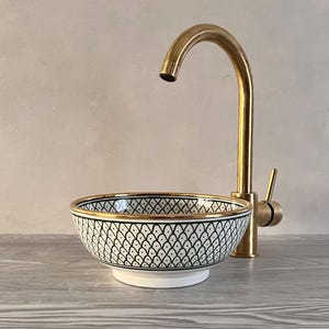 May include: A white ceramic sink with a black and white geometric pattern and a gold rim. The sink is paired with a gold faucet.