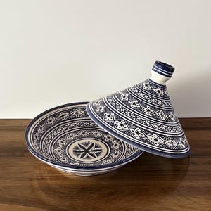 Peut inclure: Une tagine en céramique blanche avec un motif géométrique bleu et blanc. La tagine a une base ronde et un couvercle conique.