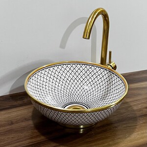 May include: A patterned ceramic sink with a gold rim and a black and white geometric design. A gold tap is installed on the side of the sink. The sink is on a dark wood surface.