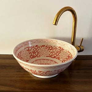 Peut inclure: Un évier décoratif en céramique avec un motif floral rouge et blanc. L'évier est rond et repose sur une surface en bois foncé. Un robinet doré est monté sur le côté.