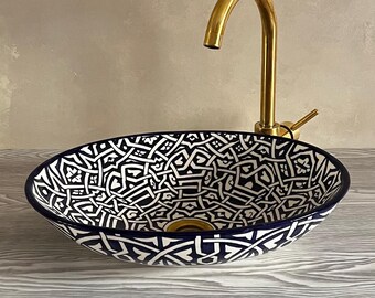 Può includere: Un lavabo da bagno in ceramica bianca con un motivo geometrico blu e bianco. Il lavabo è di forma ovale e ha un rubinetto dorato.