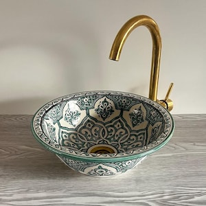Op de afbeelding: Een witte keramische wastafel met een groen-wit geometrisch patroon. De wastafel is rond en heeft een gouden kraan.