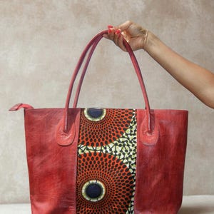 May include: A red leather tote bag with a central panel featuring a vibrant African print in orange, black, and white. The bag has two handles and a zipper closure, showcasing a blend of textures and colors.