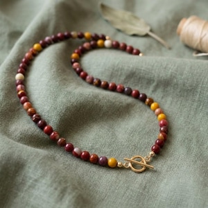 May include: A beaded necklace featuring round, multi-coloured stones in shades of deep red, maroon, brown, and yellow. The necklace is secured with a gold-toned metal toggle clasp, consisting of a circular ring and a T-bar connector piece.