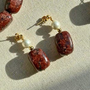 Könnte beinhalten: Ein Paar goldfarbene Ohrstecker. Jeder Ohrring hat eine weiße Perle, einen rechteckigen, rotbraunen Stein und goldfarbene Akzente. Die Ohrringe werden auf einem hellbeigen Stoff präsentiert.
