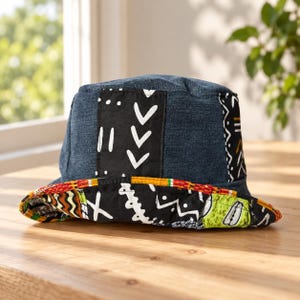 May include: A bucket hat featuring a dark blue denim crown with a vertical black panel displaying white geometric symbols. The brim is lined with a vibrant, multi-colored fabric pattern in shades of red, yellow, green, and black with white motifs.