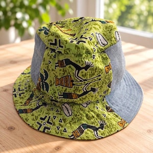 Può includere: Un cappello a secchiello in tessuto verde lime con una stampa nera e gialla di figure stilizzate e forme geometriche, a contrasto con pannelli in denim azzurro sui lati e sulla parte inferiore della tesa larga.
