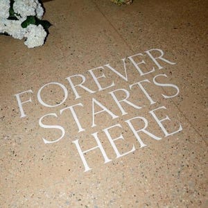 Może przedstawiać: Jasnobrązowa, nakrapiana podłoga z białym tekstem "FOREVER STARTS HERE". Tekst jest prostą, elegancką czcionką. Obraz sugeruje ślub lub specjalne wydarzenie.