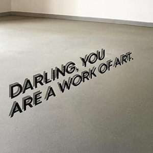 Könnte beinhalten: Minimalistischer Innenraum mit dem Text "DARLING, YOU ARE A WORK OF ART" in fetten 3D-Buchstaben auf einem hellgrauen Boden. Der Text ist abgewinkelt und erzeugt einen dynamischen visuellen Effekt vor neutralem Hintergrund.