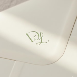 Op de afbeelding: Close-up van een crèmekleurig briefpapier met een groen monogram van de letters "D" en "L". De letters zijn in een sierlijke, cursieve lettertype. Het papier heeft een subtiele textuur en is zacht verlicht.