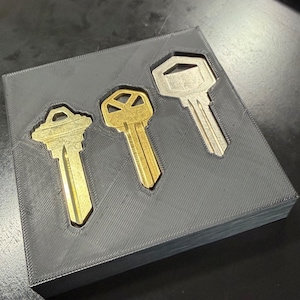 May include: A square, dark gray, 3D-printed tray featuring three recessed cutouts shaped to hold keys. Two of the cutouts contain gold-colored metal keys, while the third cutout on the right holds a silver-colored metal key. The tray has a visible diagonal layer line texture across its surface.