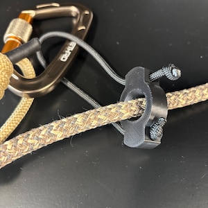 May include: Close-up of climbing gear, including a silver carabiner with an orange accent, a black rope management device, and a tan and brown climbing rope. The gear is set against a dark background.