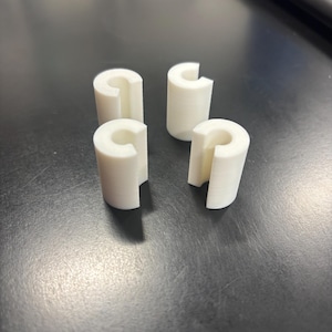 May include: Four white, C-shaped plastic components are arranged on a dark surface. Each piece has a cylindrical form with a section removed, creating the C shape. The objects appear to be 3D-printed.