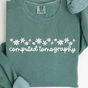 Puede incluir: Una sudadera de color verde claro con un diseño de margaritas blancas y el texto "computed tomography" impreso en la parte delantera.