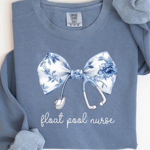 Könnte beinhalten: Blaues Jeans-Sweatshirt mit einer floralen Schleife und einer Stethoskop-Grafik. Die Schleife hat einen weißen Hintergrund mit blauen Blumenmustern. Der Text "float pool nurse" ist darunter gedruckt.