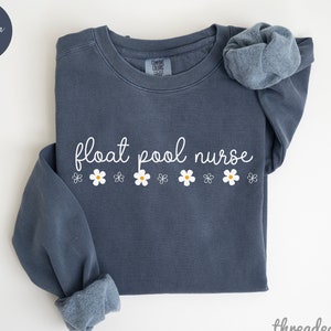 Könnte beinhalten: Ein Sweatshirt in Denim-Blau mit dem Text "float pool nurse" in weißer Kursivschrift und einer Reihe weißer Gänseblümchen mit gelben Zentren unter dem Text.