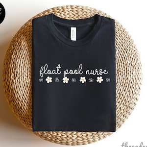 Może przedstawiać: Czarny T-shirt z napisem "float pool nurse" w białym kolorze i rzędem białych stokrotek z żółtymi środkami.
