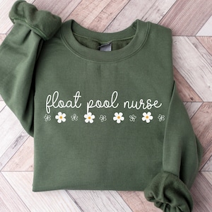 Könnte beinhalten: Ein olivgrüner Pullover mit dem Text "float pool nurse" und einer Reihe weißer Gänseblümchen mit gelben Zentren.