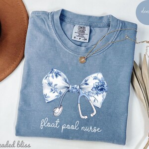 Könnte beinhalten: Ein hellblaues T-Shirt mit einer weißen und blauen Blumen Schleife und einem Stethoskop. Der Text "float pool nurse" ist auf dem Shirt gedruckt.
