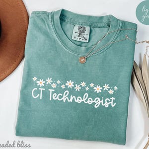 Puede incluir: Una camiseta verde claro con un borde de margaritas blancas y el texto "CT Technologist" en blanco.