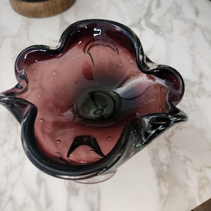 Puede incluir: Un cuenco decorativo de vidrio con borde ondulado. El cuenco es de color púrpura oscuro con un borde más oscuro y una textura de burbujas. El cuenco tiene un centro redondo de color púrpura oscuro.