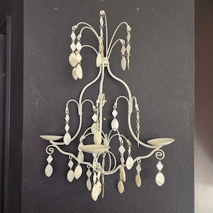 May include: Off-white metal wall sconce with a chandelier design. The sconce features two candle holders and is adorned with hanging, decorative, shell-shaped and diamond-shaped accents. The sconce is mounted on a dark gray wall.