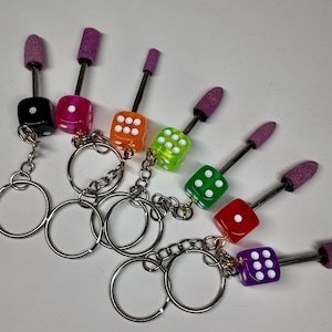 May include: A collection of colorful dice keychains. Each keychain features a die in a different color: black, pink, orange, green, red, and purple. Each die has white dots and is attached to a silver chain and ring. The keychains also have a purple grinding bit.