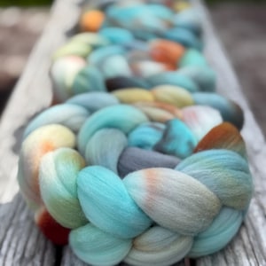 May include: A thick, braided length of roving wool featuring a variegated color palette of soft turquoise, light blue, muted grey, cream, and accents of warm orange and rust, arranged in a continuous coil pattern.