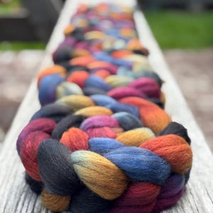 May include: A thick, braided length of roving wool featuring a variegated color palette of deep black, vibrant orange, rich burgundy, and muted blue tones. The fiber is densely packed and twisted into a continuous, textured rope-like structure.