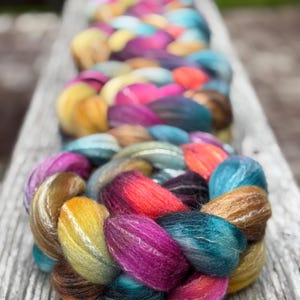 May include: A thick, braided roving of wool fiber featuring a vibrant, multi-colored gradient. The colors transition through shades of deep magenta, bright orange, golden yellow, teal, and dark blue, creating a textured, soft appearance.